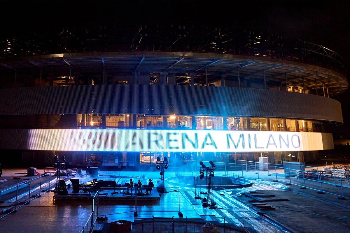 Arena Milano illuminata di notte con lavori in corso e un grande display luminoso sulla facciata.