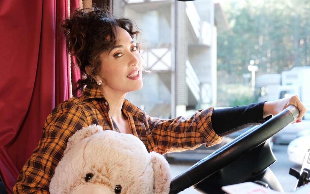 Donna sorridente guida un veicolo tenendo un grande orsacchiotto di peluche, illuminata dalla luce naturale del giorno