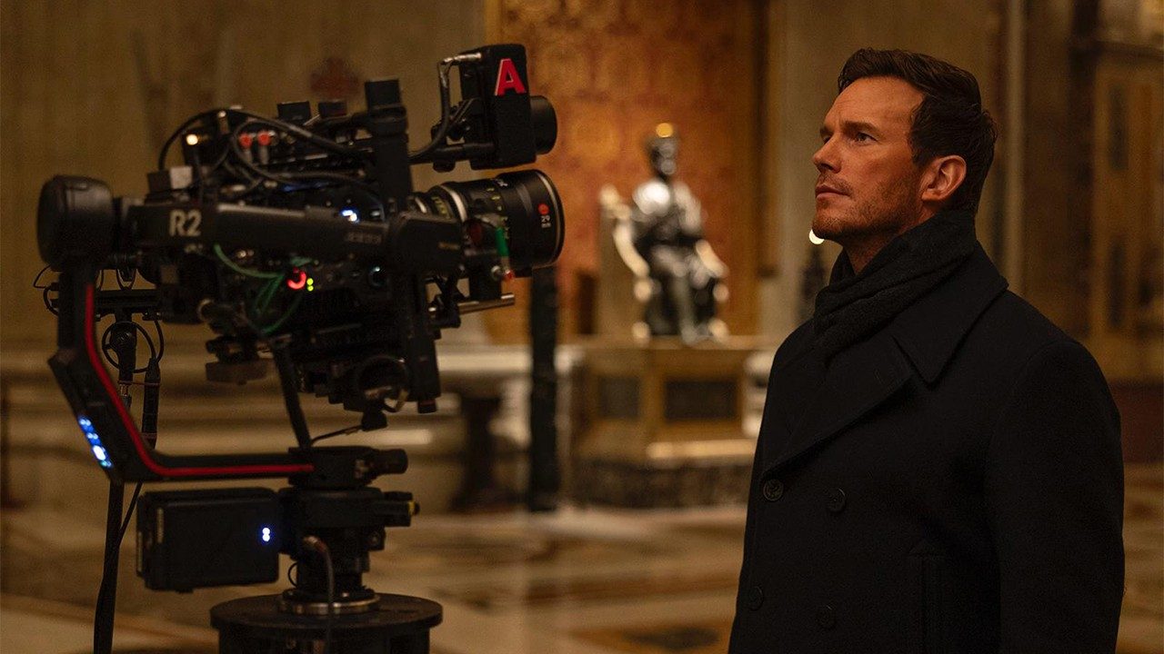 Attore di fronte a una cinepresa professionale mentre osserva una scena girata in un ambiente sacro con statua sullo sfondo.