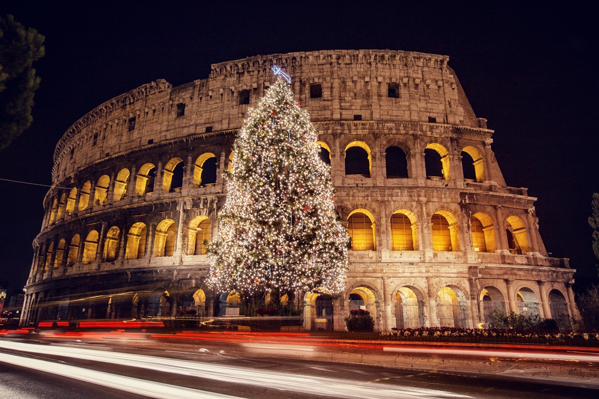 Albero di Natale illuminato davanti al Colosseo di Roma durante la notte, con luci festive e traffico in movimento.