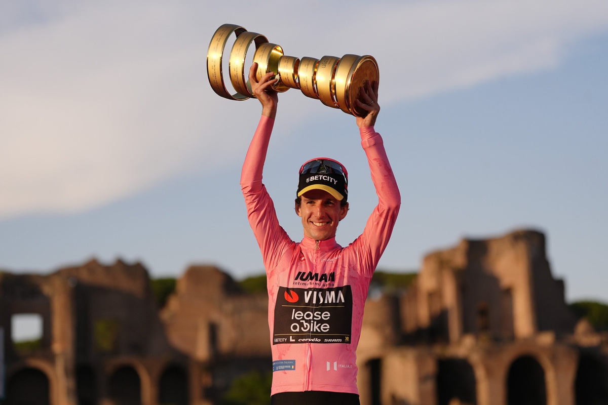 Ciclista con maglia rosa che alza un trofeo dorato davanti a rovine storiche