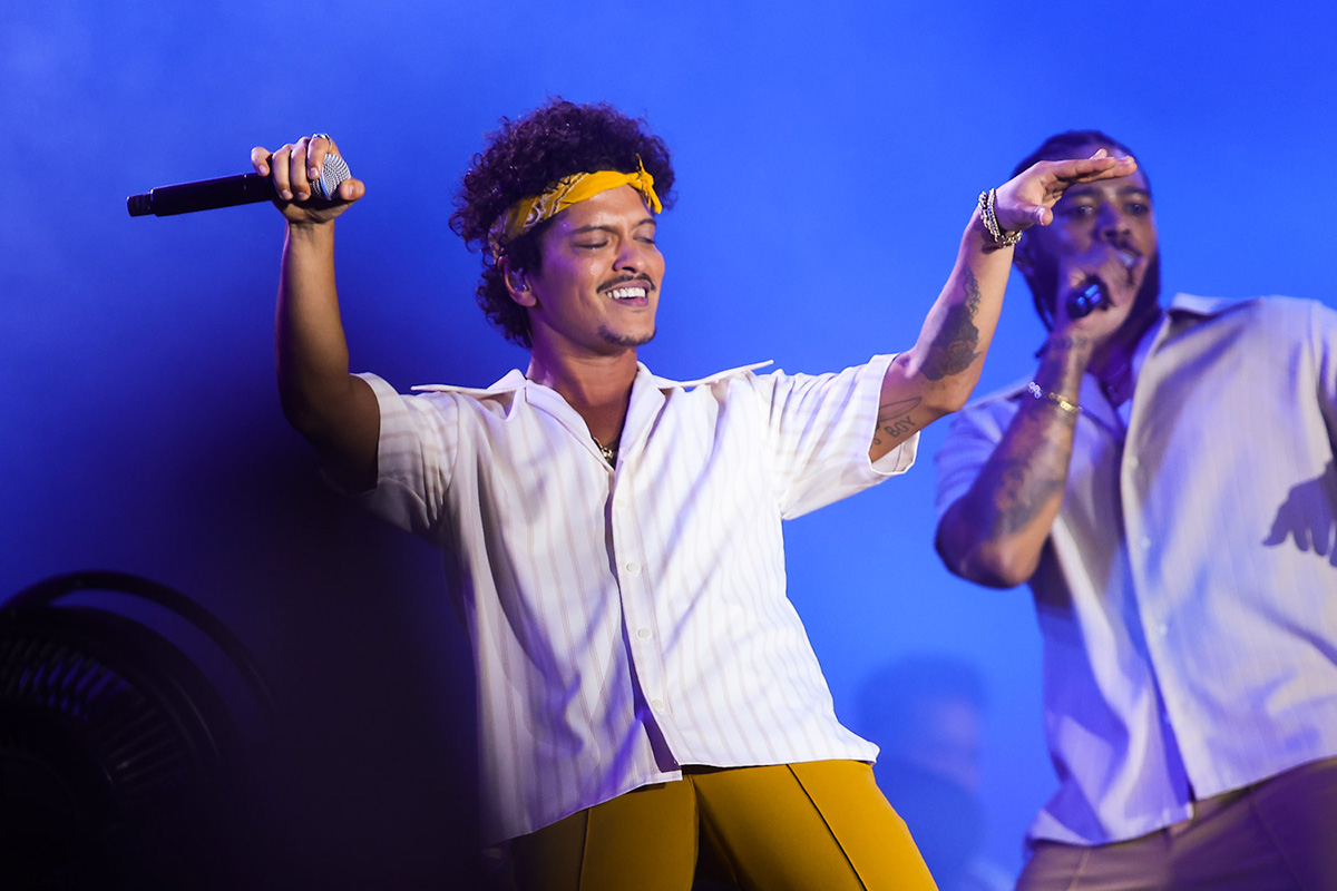 Bruno Mars canta dal vivo sul palco con microfono in mano, indossando una camicia bianca e una bandana gialla durante un concerto