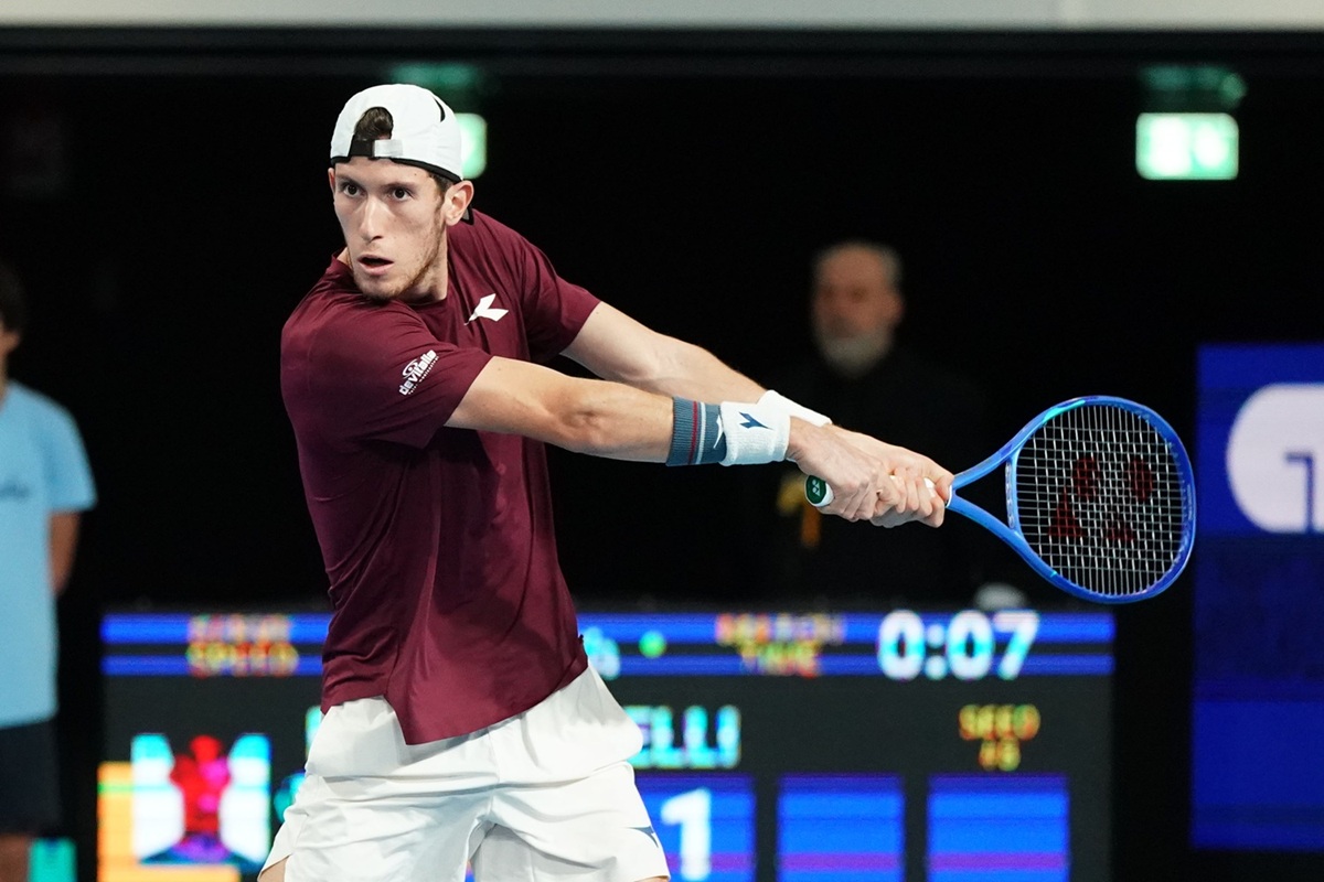 Giocatore di tennis professionista mentre esegue un rovescio a due mani in un torneo indoor, con racchetta blu e abbigliamento sportivo bordeaux.