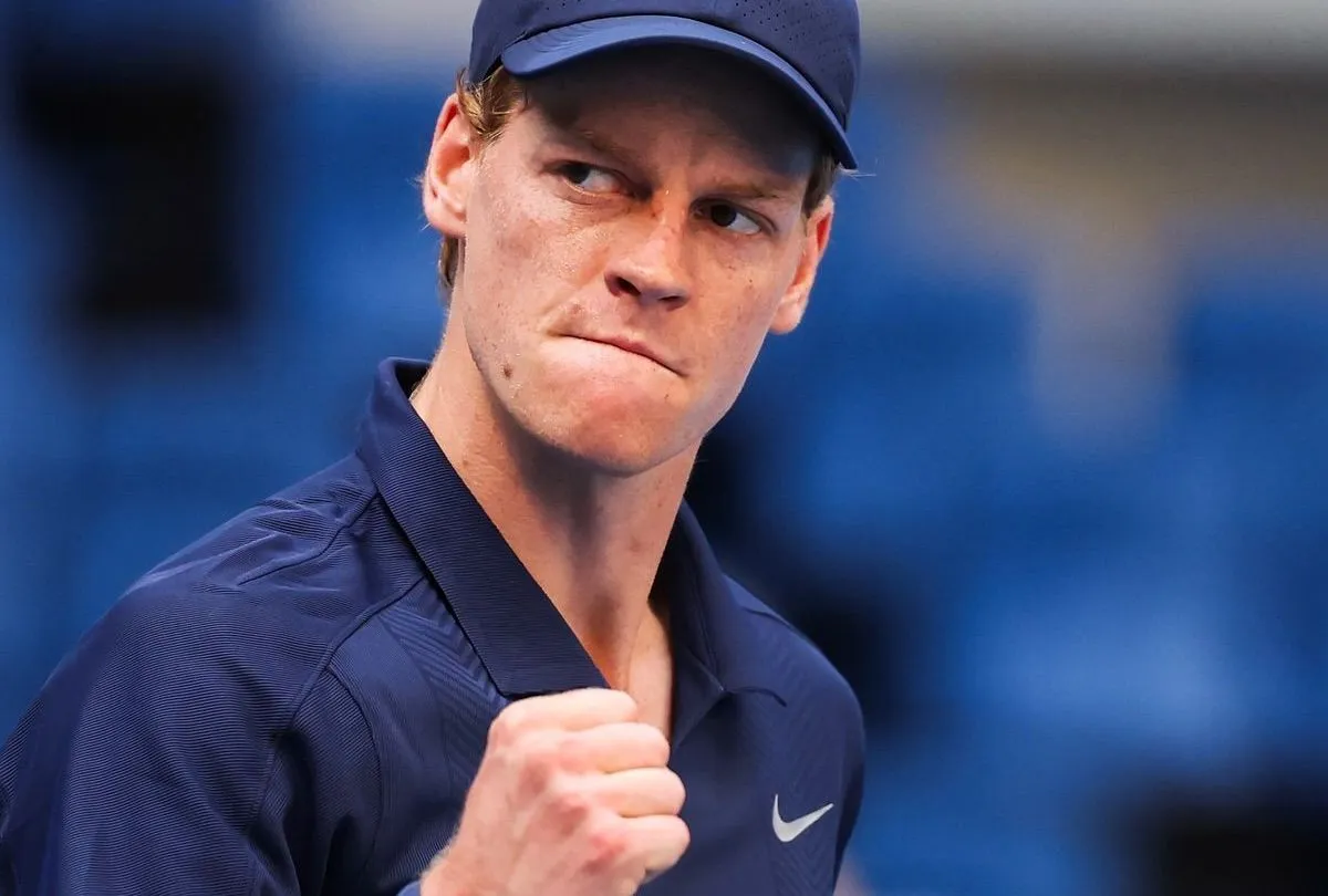 Jannik Sinner in primo piano durante una partita di tennis, con pugno chiuso e sguardo concentrato, indossa cappellino e maglia blu.