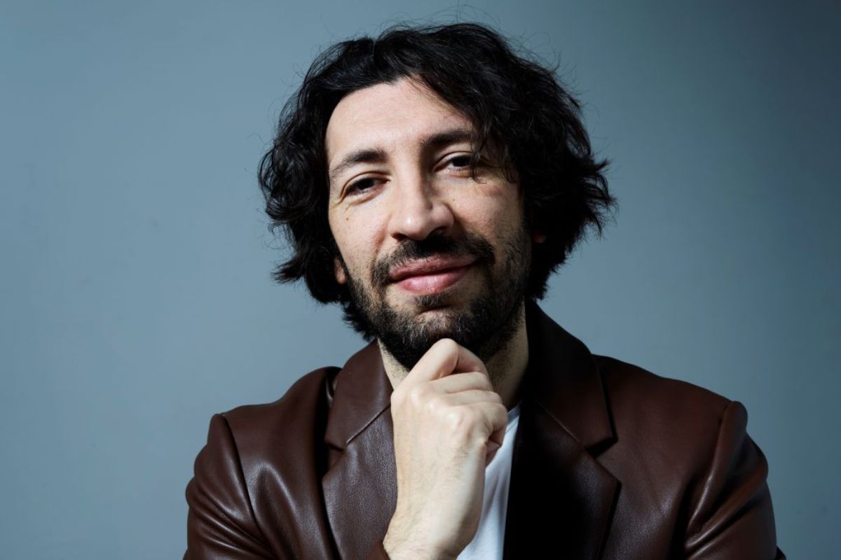 Uomo con barba e capelli ricci scuri, ritratto in primo piano, posa pensierosa con mano sul mento e giacca marrone.