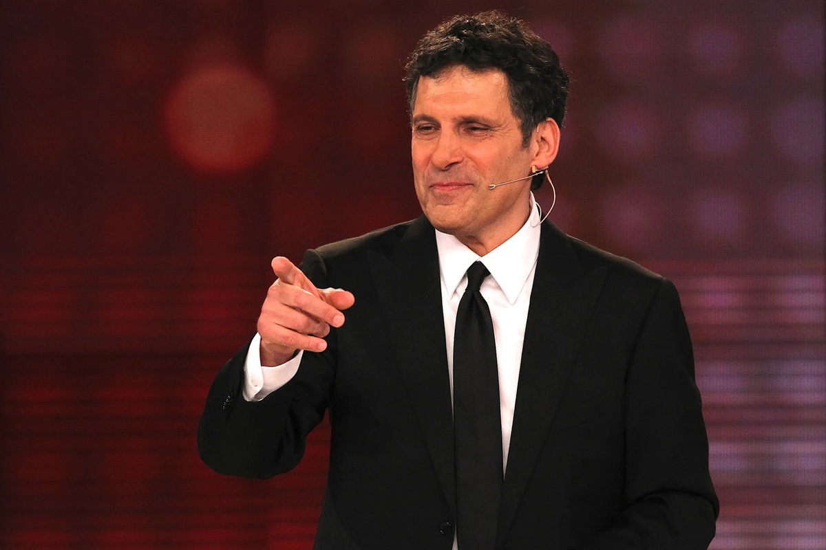 Conduttore televisivo in abito nero che parla al pubblico durante uno show televisivo in studio, con microfono auricolare e sfondo luminoso