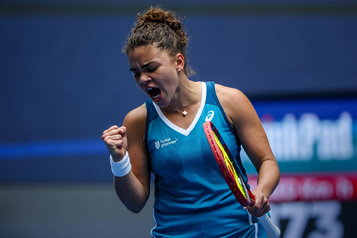 Tennista donna che esulta durante una partita, con racchetta in mano e pugno chiuso, su un campo da tennis indoor.