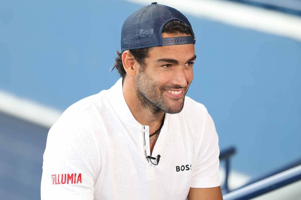 Tennista professionista con cappellino blu e maglia bianca sorride durante un’intervista su un campo da tennis in cemento.