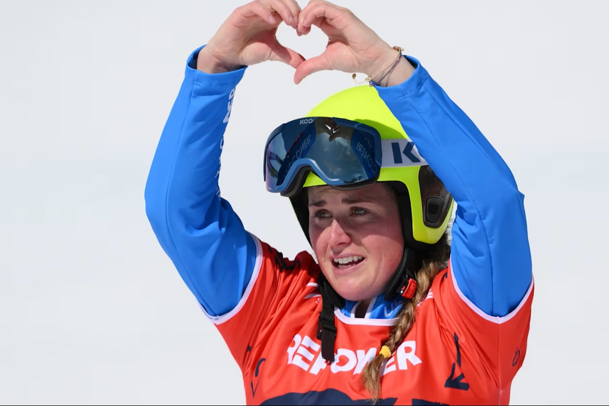 Atleta di snowboard con casco e occhiali forma un cuore con le mani durante una competizione sulla neve.