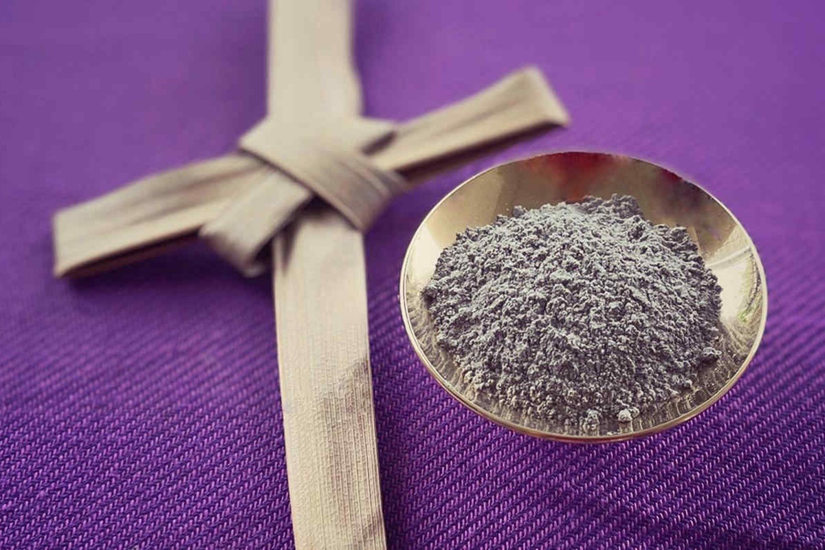 Ceneri del Mercoledì delle Ceneri in una ciotola metallica accanto a una croce intrecciata di palma su tessuto viola