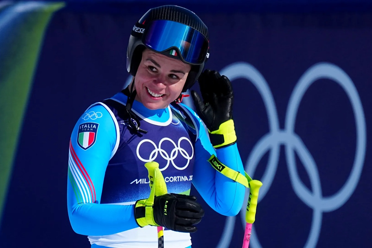 Sciatrice del Team Italia sorride durante una gara alle Olimpiadi Invernali Milano Cortina 2026 con casco, occhiali e bastoncini da sci.