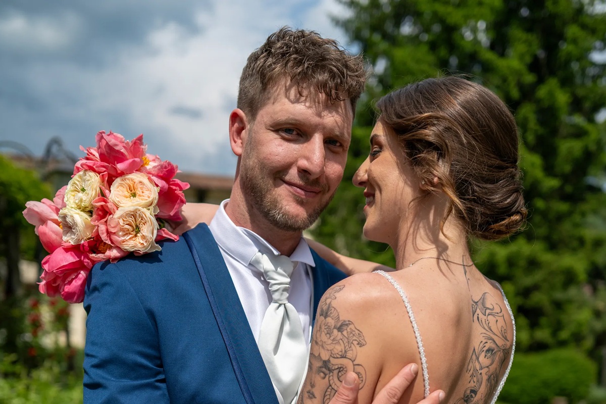 Coppia di sposi durante matrimonio all’aperto, lo sposo con bouquet di rose e la sposa con abito bianco elegante.