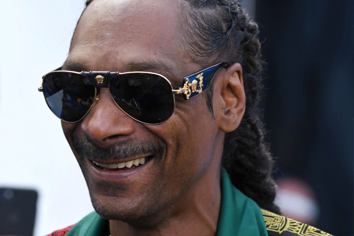 Uomo sorridente con occhiali da sole di lusso, capelli intrecciati e abbigliamento elegante durante un evento all’aperto.