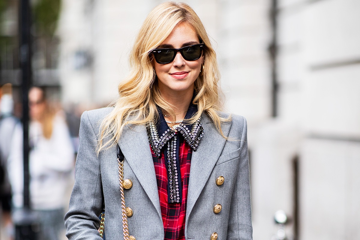 Influencer di moda con capelli biondi, occhiali da sole e blazer grigio elegante mentre cammina in una strada della città