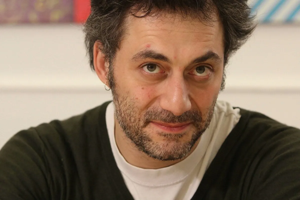 Un uomo con la barba corta e i capelli scuri guarda in camera in un ambiente interno con uno sfondo sfocato.