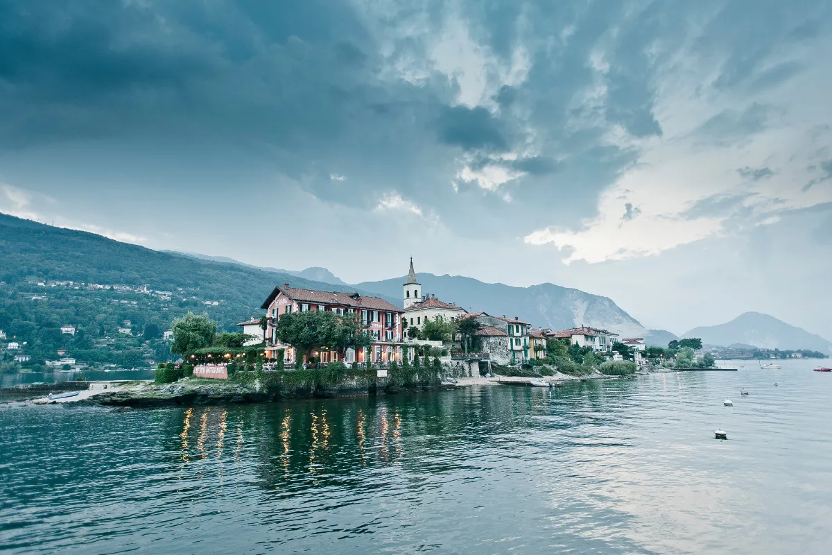 Un piccolo villaggio con case colorate e una chiesa su un'isola del Lago Maggiore, circondato da montagne e acque calme.