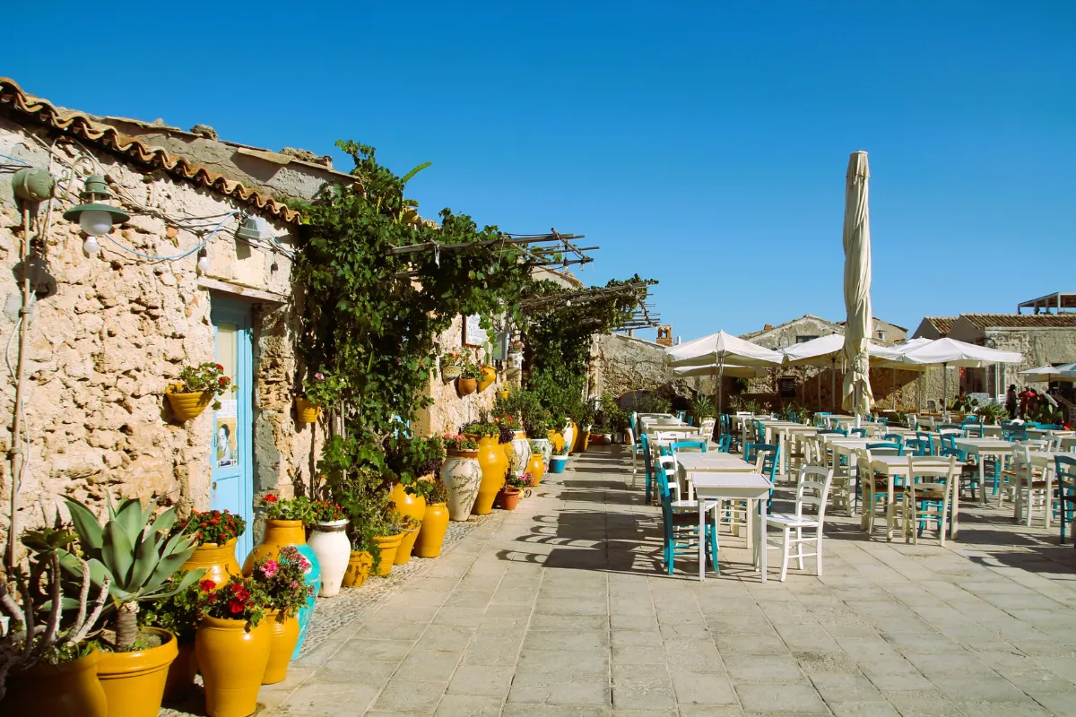 Una piazza con tavoli da ristorante all'aperto accanto a case in pietra decorate con piante in vaso a Marzamemi, in Sicilia.