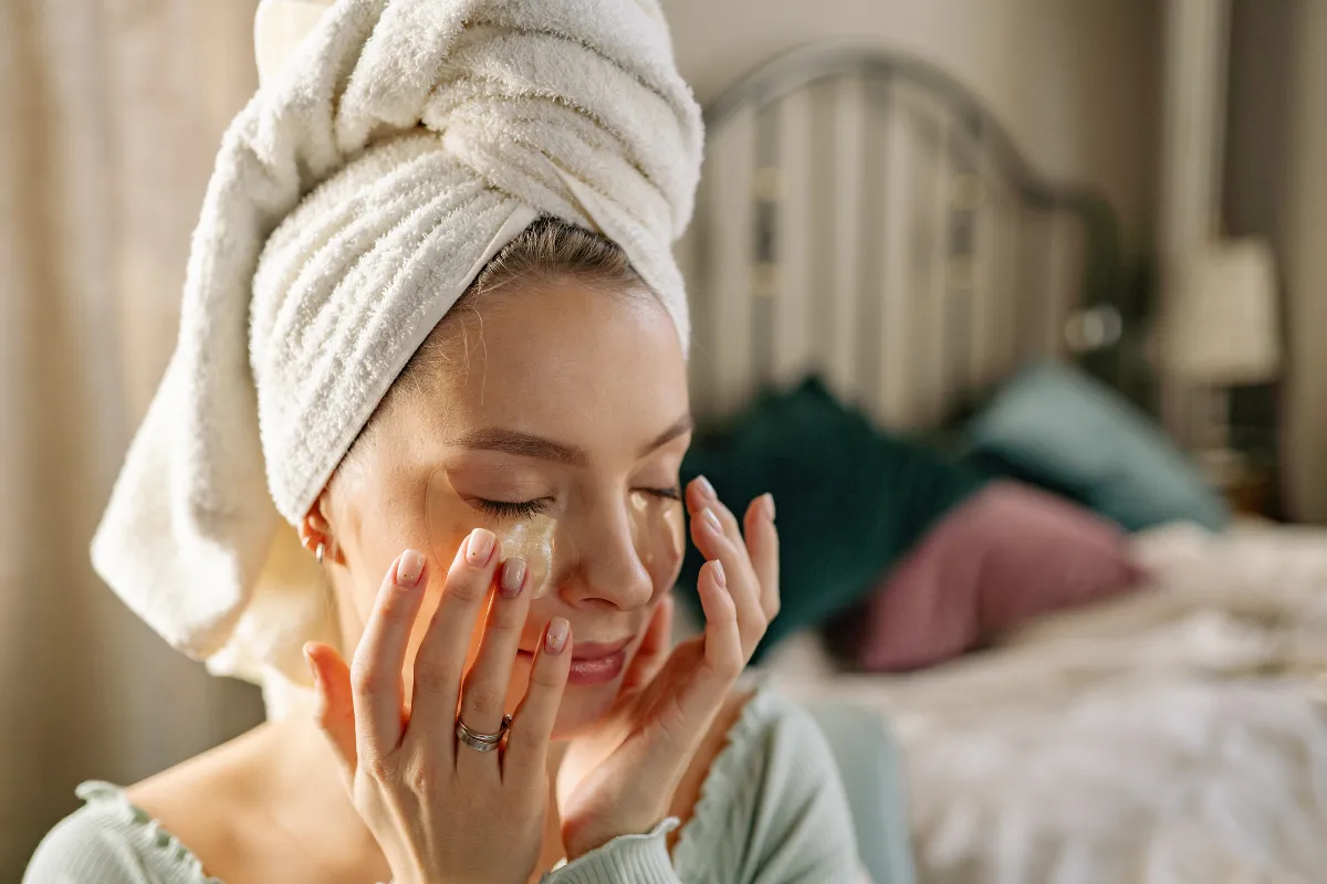 Una donna con un asciugamano sulla testa si applica una maschera per gli occhi davanti a uno specchio.