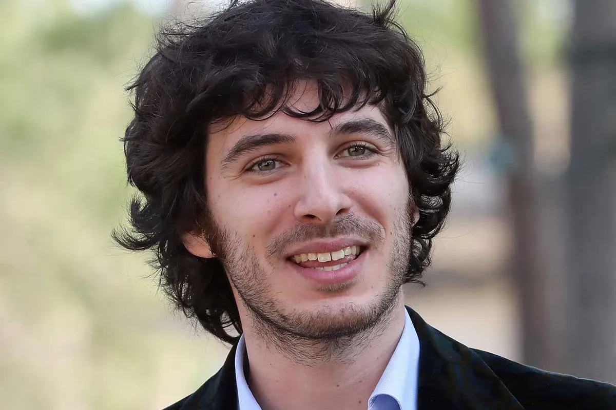 Giovane uomo con capelli ricci scuri e barba leggera che sorride all’aperto con sfondo naturale sfocato