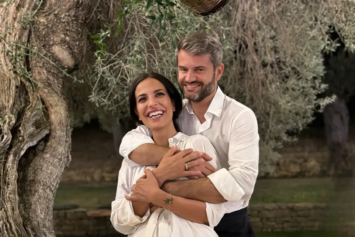 Coppia elegante vestita di bianco abbracciata sotto un ulivo in un giardino, sorridente e felice durante un momento romantico all’aperto.