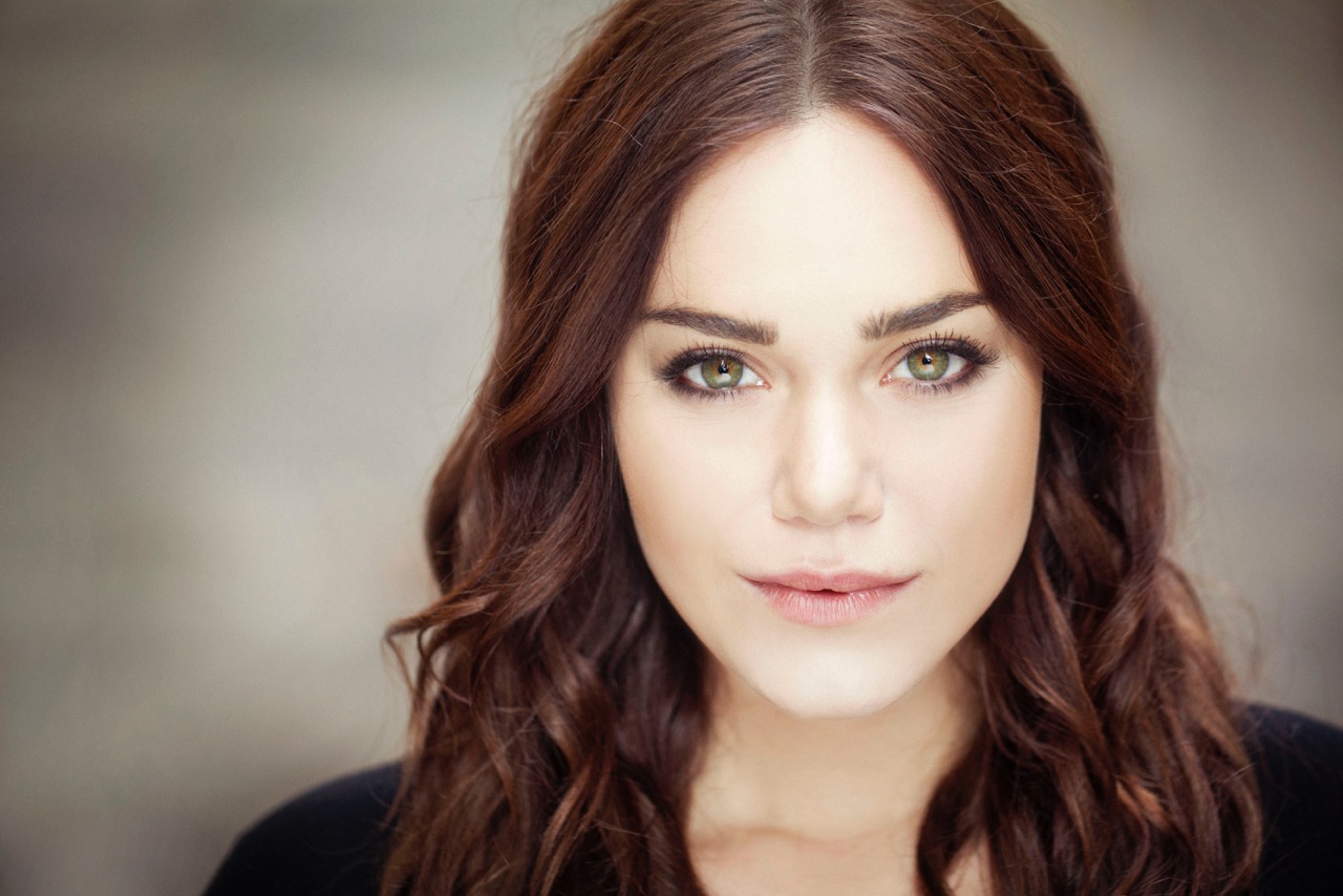 Primo piano di donna con occhi verdi intensi e capelli castani mossi, sfondo neutro sfocato.