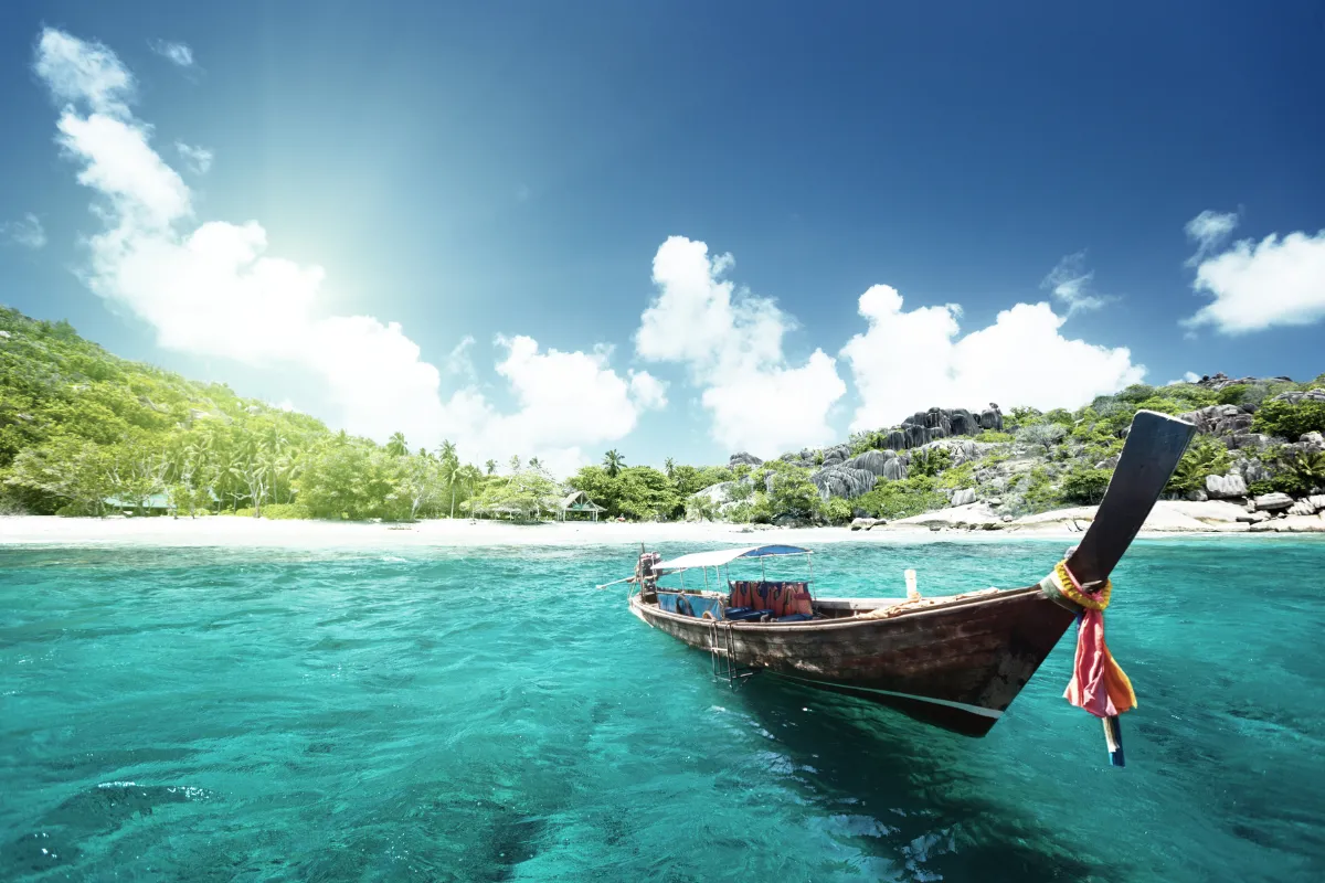 Una tradizionale barca di legno che naviga su acque cristalline vicino a una spiaggia con vegetazione tropicale e cielo azzurro in Thailandia.
