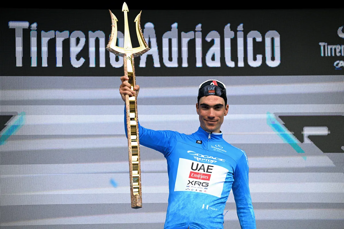 Ciclista vincitore della Tirreno-Adriatico 2026 con maglia azzurra che solleva il tridente dorato sul podio