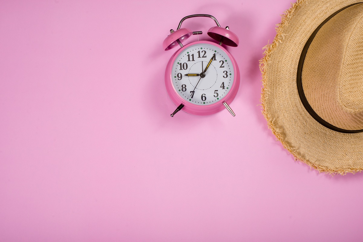 Orologio sveglia rosa accanto a un cappello di paglia su sfondo rosa pastello minimalista