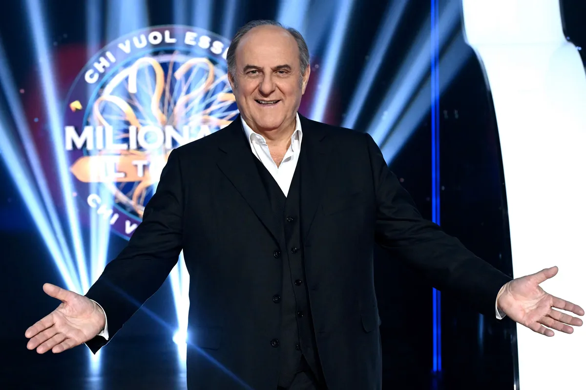 Conduttore televisivo sorridente sul palco di un quiz show con luci scenografiche e logo Chi Vuol Essere Milionario sullo sfondo