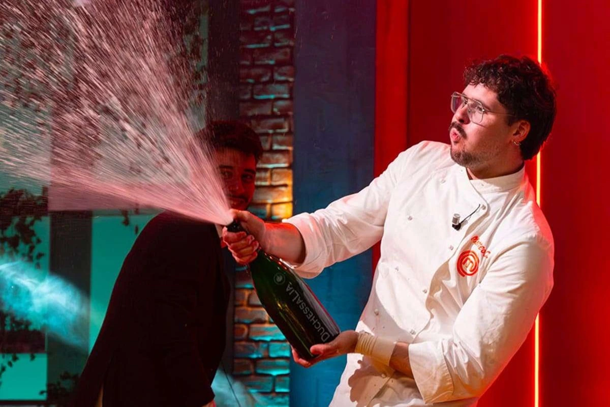 Chef con giacca da cucina di MasterChef Italia apre una bottiglia di champagne spruzzando durante una celebrazione in studio