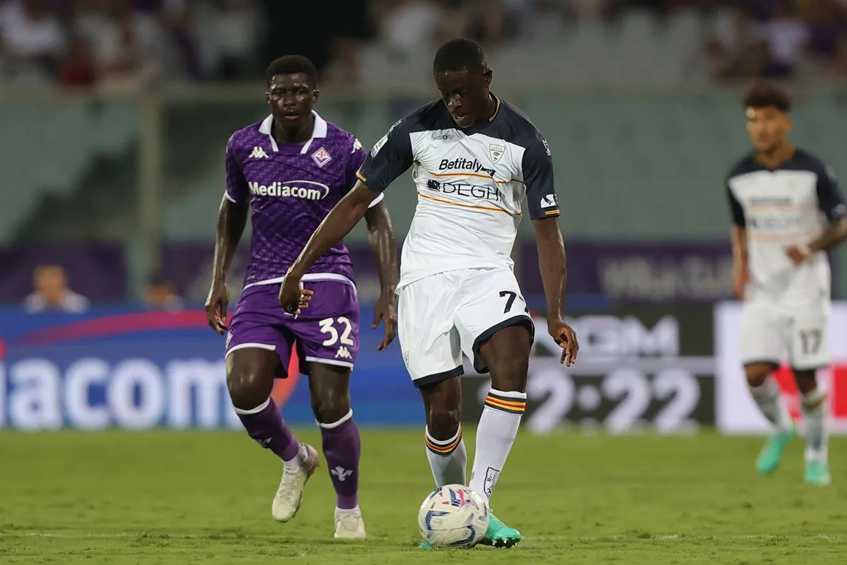 Giocatore del Lecce in maglia bianca dribbla il pallone mentre un avversario della Fiorentina insegue durante una partita di calcio