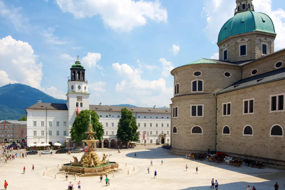 Una grande piazza a Salisburgo con una fontana centrale, edifici storici, una cattedrale con una cupola verde e gente che passeggia.